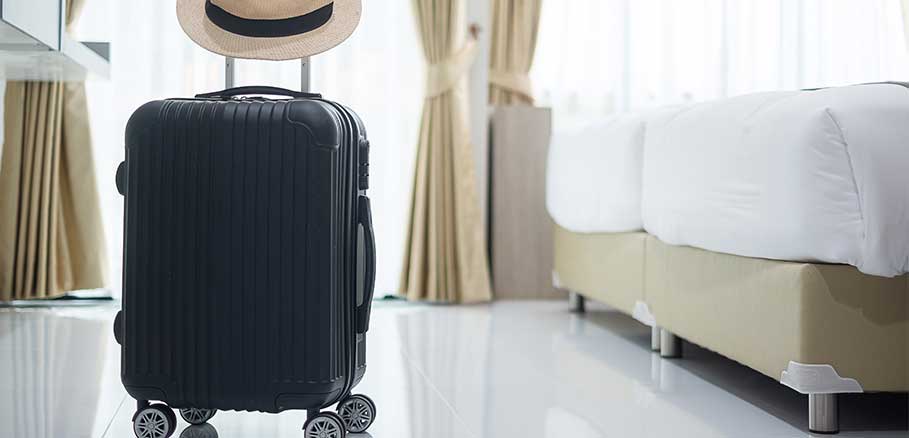 Μαύρη βαλίτσα με ρόδες και ψάθινο καπέλο σε δωμάτιο ξενοδοχείου